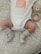 A baby wearing a white onesie with a peach design and the words 'PEACHY KEEN' written on it. The baby has gray socks with red text that reads 'MY FIRST XMAS'. A knitted teddy bear rattle is next to the baby, resting on a patterned surface.