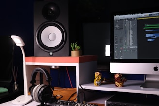 A well-organized workspace featuring an audio studio setup including a large speaker, a modern desktop computer displaying audio editing software, a pair of headphones resting on a stand, and a small potted plant for decor. The setup is illuminated by a sleek desk lamp, creating a warm, inviting atmosphere. Decorative owl figures also add a touch of personality to the space.