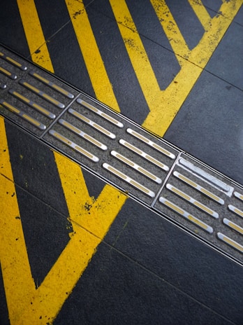 A textured metal strip runs diagonally across a dark surface with prominent yellow chevron markings. The surface appears worn and industrial, suggesting an area intended for guiding or directing movement.