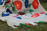 A piece of fabric with a floral design featuring vibrant red and blue flowers connected by green stems and leaves. The fabric is draped over grass, creating a natural backdrop that contrasts with the vivid colors of the textile.