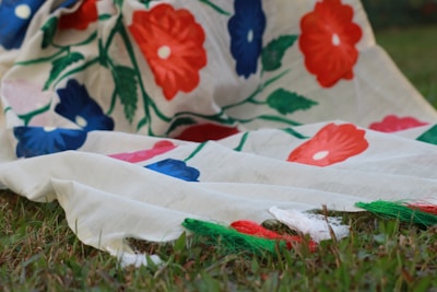 A piece of fabric with a floral design featuring vibrant red and blue flowers connected by green stems and leaves. The fabric is draped over grass, creating a natural backdrop that contrasts with the vivid colors of the textile.