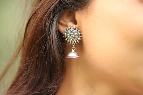 An ornate silver earring with a sunburst design is worn by a person. The earring features small intricate details and hangs delicately from the earlobe. The background is a soft color, focusing attention on the jewelry.