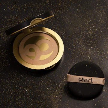 A compact face powder in a circular black and gold container, opened to reveal the powder inside with a design resembling a Cancer zodiac symbol. A black applicator sponge with a beige ribbon labeled 'Berl' is placed beside it. The background is a textured black surface with scattered gold specks.