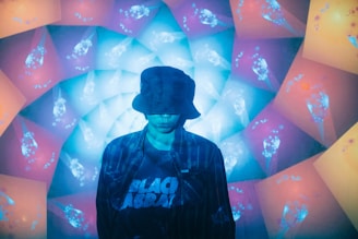A person wearing a bucket hat stands in front of a vibrant, kaleidoscopic backdrop composed of blue, purple, and orange glowing patterns. The person wears a shirt with the text 'Black Sabbath'. The overall lighting casts a blue hue on the person, creating a surreal effect.