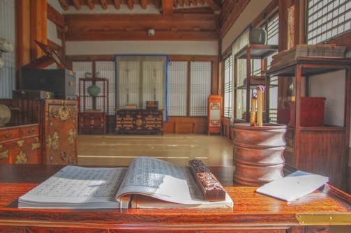 A serene, traditional interior space features wooden beams and intricate furniture. An open book with music sheets is placed on a polished wooden table, accompanied by a carved pen or writing tool. Sunlight filters through the lattice windows, casting soft shadows and highlighting the room's carefully arranged artifacts and decorative elements.