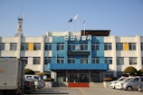 A three-story building with a blue and white facade, featuring a sign with Korean text at the top. Several flags are flying above the building. There is a telecommunications tower to the left. In front of the building, multiple vehicles, including police cars, are parked, and there are neatly trimmed bushes lining the parking area.