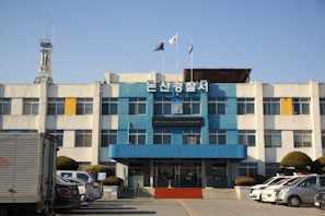 A three-story building with a blue and white facade, featuring a sign with Korean text at the top. Several flags are flying above the building. There is a telecommunications tower to the left. In front of the building, multiple vehicles, including police cars, are parked, and there are neatly trimmed bushes lining the parking area.
