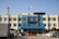 A three-story building with a blue and white facade, featuring a sign with Korean text at the top. Several flags are flying above the building. There is a telecommunications tower to the left. In front of the building, multiple vehicles, including police cars, are parked, and there are neatly trimmed bushes lining the parking area.