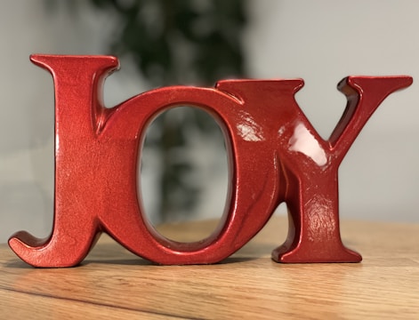 Shiny red ceramic letters spelling out the word 'JOY' are placed on a wooden surface. The background is softly blurred with hints of green, possibly from a plant.