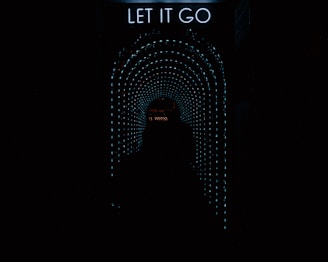 A dark tunnel with bright, dotted light patterns forming a circular arch converging towards the center. At the top of the image, there is a neon sign that says 'LET IT GO.' A silhouette of a person is standing in the tunnel, facing the lights