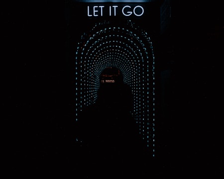 A dark tunnel with bright, dotted light patterns forming a circular arch converging towards the center. At the top of the image, there is a neon sign that says 'LET IT GO.' A silhouette of a person is standing in the tunnel, facing the lights
