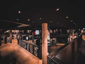 A dimly lit, modern library interior with multiple levels and extensive bookshelves lining the walls. The space features a mix of industrial and contemporary design elements, including concrete pillars and metal railings. The warm lighting casts soft shadows, creating an intimate atmosphere.