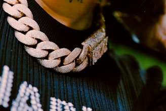 A close-up view of a thick, gold chain necklace with a large clasp, adorned with sparkling stones or diamonds. The background shows part of a dark, ribbed fabric.