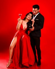 A woman in a flowing red dress with a thigh-high slit is holding a glass of red wine. She is laughing and leaning back slightly against a man in a black suit. The background is a vivid red, matching the woman's dress and the energetic atmosphere.
