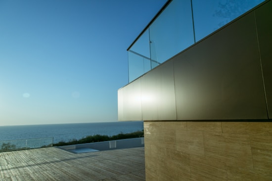 A modern building with a glass facade overlooks a serene ocean view. The sunlight creates a soft reflection on the building's surface. A wide wooden deck stretches towards the edge, with a glimpse of greenery and the horizon.