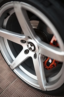 Close-up of a car wheel featuring a silver alloy rim, with the BMW logo at the center. The wheel has a sporty design with five spokes and visible brake caliper, which is painted in orange.