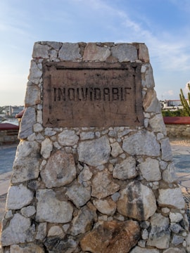 A stone monument made of irregularly shaped rocks with a plaque bearing the word 'INOLVIDABLE'. The background shows a cityscape with buildings under a clear blue sky.