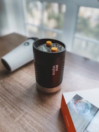 A Godox ML60 light rests on a wooden surface with two yellow dials on top. Nearby, there is a white cylindrical object and a product box featuring an image with a color wheel. The background is softly blurred, indicating a window with natural light coming through.