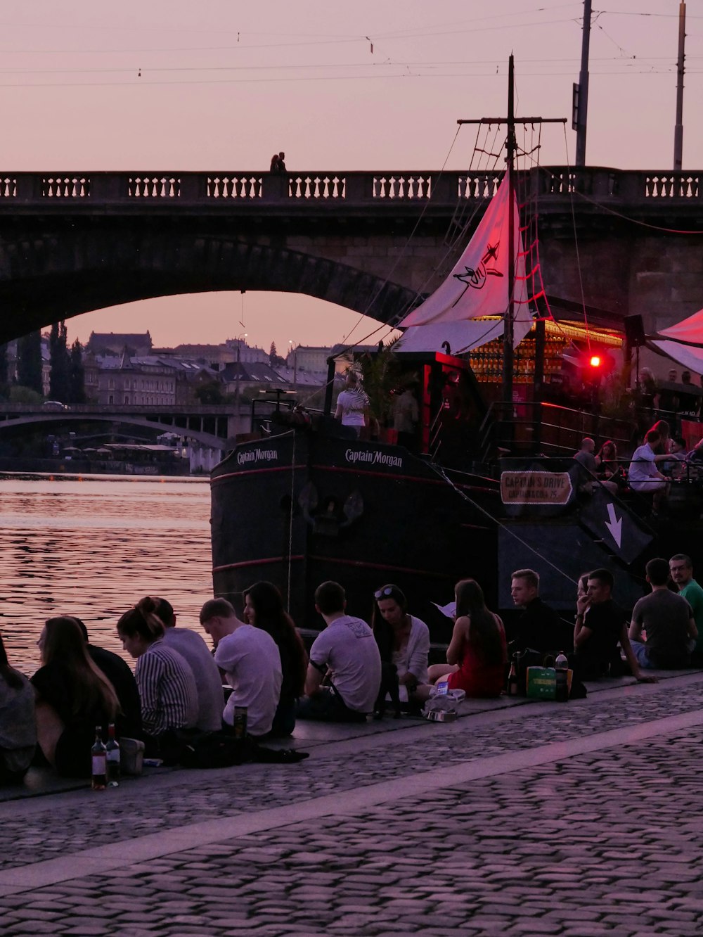 a group of people sitting next to a river under a bridge