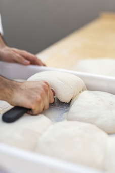 A hand is using a dough scraper to divide a large batch of unbaked dough into smaller portions. The dough is white and fluffy, resting in a rectangular white container. The background is a light wooden surface.