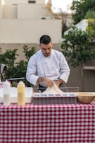 A person in a white chef's jacket is preparing food at an outdoor table covered with a red and white checkered tablecloth. The table has several items, including condiment bottles and a wooden bowl. Lush greenery and beige buildings are in the background.