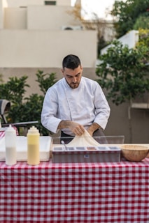 A person in a white chef's jacket is preparing food at an outdoor table covered with a red and white checkered tablecloth. The table has several items, including condiment bottles and a wooden bowl. Lush greenery and beige buildings are in the background.