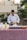 A person in a white chef's jacket is preparing food at an outdoor table covered with a red and white checkered tablecloth. The table has several items, including condiment bottles and a wooden bowl. Lush greenery and beige buildings are in the background.