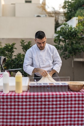 A person in a white chef's jacket is preparing food at an outdoor table covered with a red and white checkered tablecloth. The table has several items, including condiment bottles and a wooden bowl. Lush greenery and beige buildings are in the background.