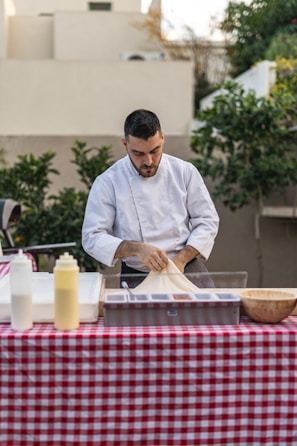 A person in a white chef's jacket is preparing food at an outdoor table covered with a red and white checkered tablecloth. The table has several items, including condiment bottles and a wooden bowl. Lush greenery and beige buildings are in the background.