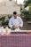 A person in a white chef's jacket is preparing food at an outdoor table covered with a red and white checkered tablecloth. The table has several items, including condiment bottles and a wooden bowl. Lush greenery and beige buildings are in the background.