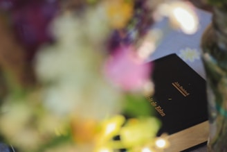A book with a dark cover and gold lettering is partially visible among blurred flowers. The title and author's name appear on the book cover.
