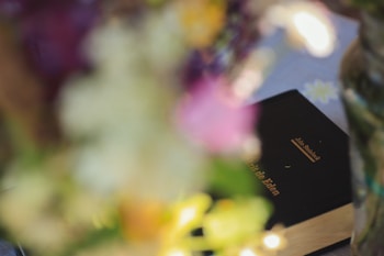 A book with a dark cover and gold lettering is partially visible among blurred flowers. The title and author's name appear on the book cover.