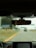A view from inside a car focusing on the rearview mirror, which reflects the driver's eyes. The road ahead is visible through the windshield, slightly blurred, with some road signs in the distance. The car dashboard is partially visible at the bottom.