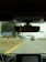 A view from inside a car focusing on the rearview mirror, which reflects the driver's eyes. The road ahead is visible through the windshield, slightly blurred, with some road signs in the distance. The car dashboard is partially visible at the bottom.