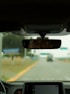 A view from inside a car focusing on the rearview mirror, which reflects the driver's eyes. The road ahead is visible through the windshield, slightly blurred, with some road signs in the distance. The car dashboard is partially visible at the bottom.