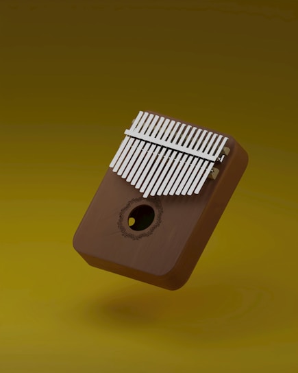 A wooden musical instrument with metal tines, known as a kalimba, is suspended against a plain mustard yellow background. The flat rectangular shape has a sound hole with a delicate decorative pattern around it.