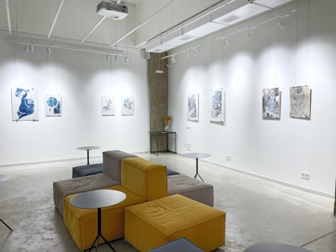 A modern gallery space featuring abstract artwork displayed on white walls under bright lighting. The room includes modular seating with yellow and gray cushions, complemented by small round tables. The ceiling has exposed fixtures and a projector.