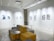 A modern gallery space featuring abstract artwork displayed on white walls under bright lighting. The room includes modular seating with yellow and gray cushions, complemented by small round tables. The ceiling has exposed fixtures and a projector.