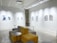A modern gallery space featuring abstract artwork displayed on white walls under bright lighting. The room includes modular seating with yellow and gray cushions, complemented by small round tables. The ceiling has exposed fixtures and a projector.