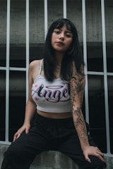 A woman with long dark hair, wearing a tank top with the word 'Angel' printed on it, is leaning against a metal fence. She has tattoos on her right arm and is dressed in casual black pants.