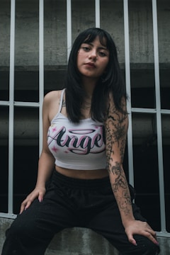 A woman with long dark hair, wearing a tank top with the word 'Angel' printed on it, is leaning against a metal fence. She has tattoos on her right arm and is dressed in casual black pants.