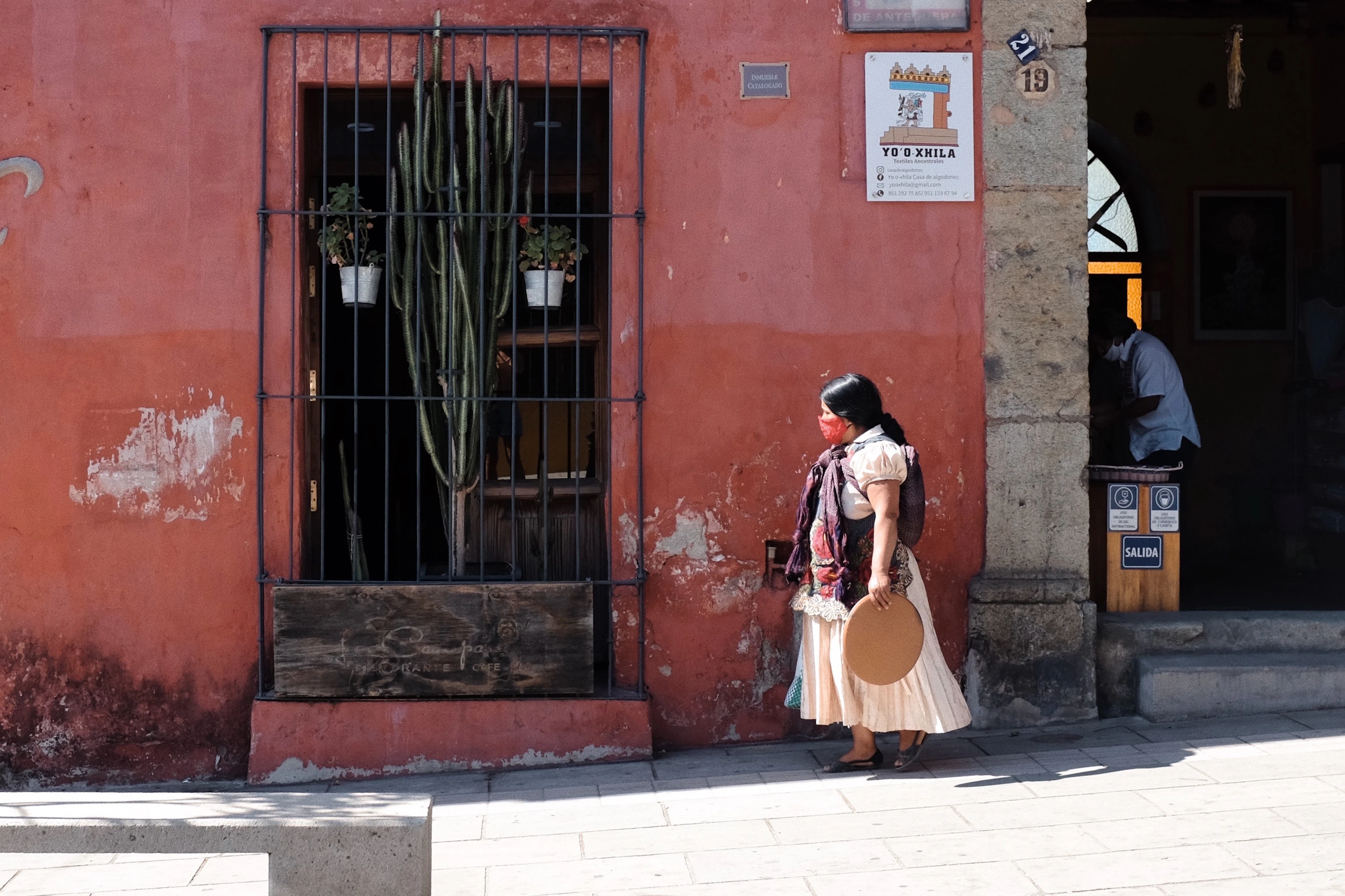 A woman in traditional Oaxacan dress walking past a weathered terracotta-red wall with a barred window and potted cactus