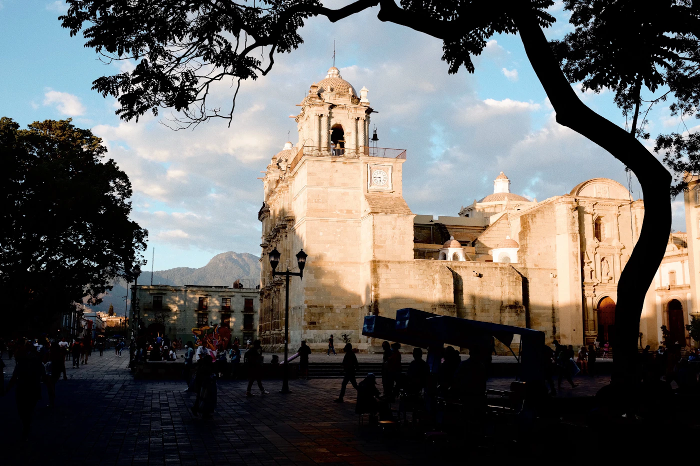 Oaxaca cathedral with clock tower photographed in warm golden afternoon light with people in the main plaza below and mountains visible in the background