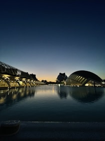 A stunning modern architecture landscape at dusk, featuring futuristic buildings with glass and metal structures reflecting on a calm water body. The sky transitions from a deep blue at the top to a warm orange near the horizon, marking the time just after sunset.