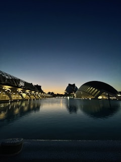 A stunning modern architecture landscape at dusk, featuring futuristic buildings with glass and metal structures reflecting on a calm water body. The sky transitions from a deep blue at the top to a warm orange near the horizon, marking the time just after sunset.