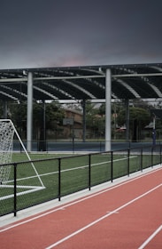 An outdoor sports facility featuring a running track and a soccer goal. The track is red, surrounded by a black metal fence, and the field is covered with artificial green turf. In the background, a basketball court is visible under a covered roof with metal supports. The sky is overcast, adding a moody atmosphere.