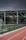 An outdoor sports facility featuring a running track and a soccer goal. The track is red, surrounded by a black metal fence, and the field is covered with artificial green turf. In the background, a basketball court is visible under a covered roof with metal supports. The sky is overcast, adding a moody atmosphere.