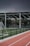 An outdoor sports facility featuring a running track and a soccer goal. The track is red, surrounded by a black metal fence, and the field is covered with artificial green turf. In the background, a basketball court is visible under a covered roof with metal supports. The sky is overcast, adding a moody atmosphere.