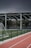 An outdoor sports facility featuring a running track and a soccer goal. The track is red, surrounded by a black metal fence, and the field is covered with artificial green turf. In the background, a basketball court is visible under a covered roof with metal supports. The sky is overcast, adding a moody atmosphere.