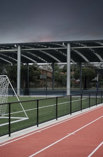 An outdoor sports facility featuring a running track and a soccer goal. The track is red, surrounded by a black metal fence, and the field is covered with artificial green turf. In the background, a basketball court is visible under a covered roof with metal supports. The sky is overcast, adding a moody atmosphere.
